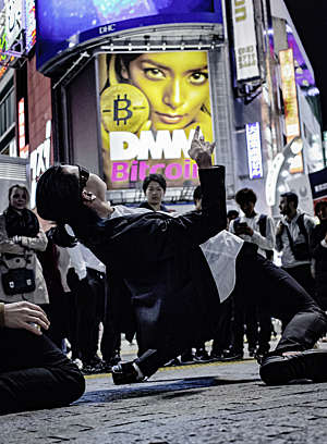 Japanese man performs a limbo dance on the street at Tokyo's Shibuya Crossing in front of a crowd of onlookers, with a big ad for Bitcoin in the background.