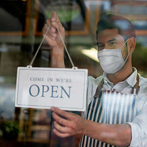 Close-up of a young man with face mask hanging an open sign on the door of a grocery store.
