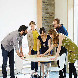 Five colleagues pore over a workplan displayed on a small round table during an animated brainstorming session.